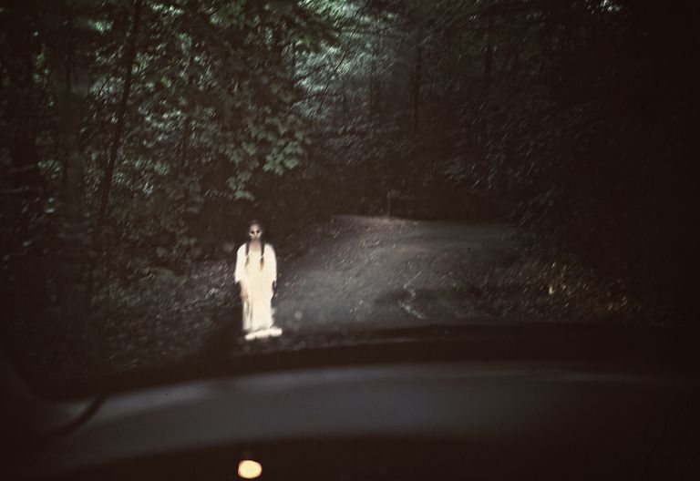 A pale figure caught in headlights on a forest road